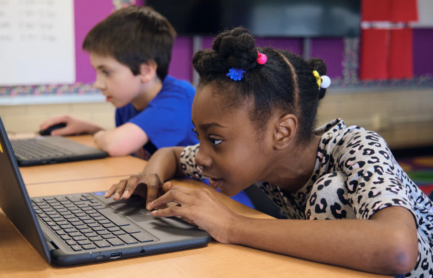 Young girl with colorful hair accessories and boy in blue shirt focused on using laptops in classroom with colorful walls